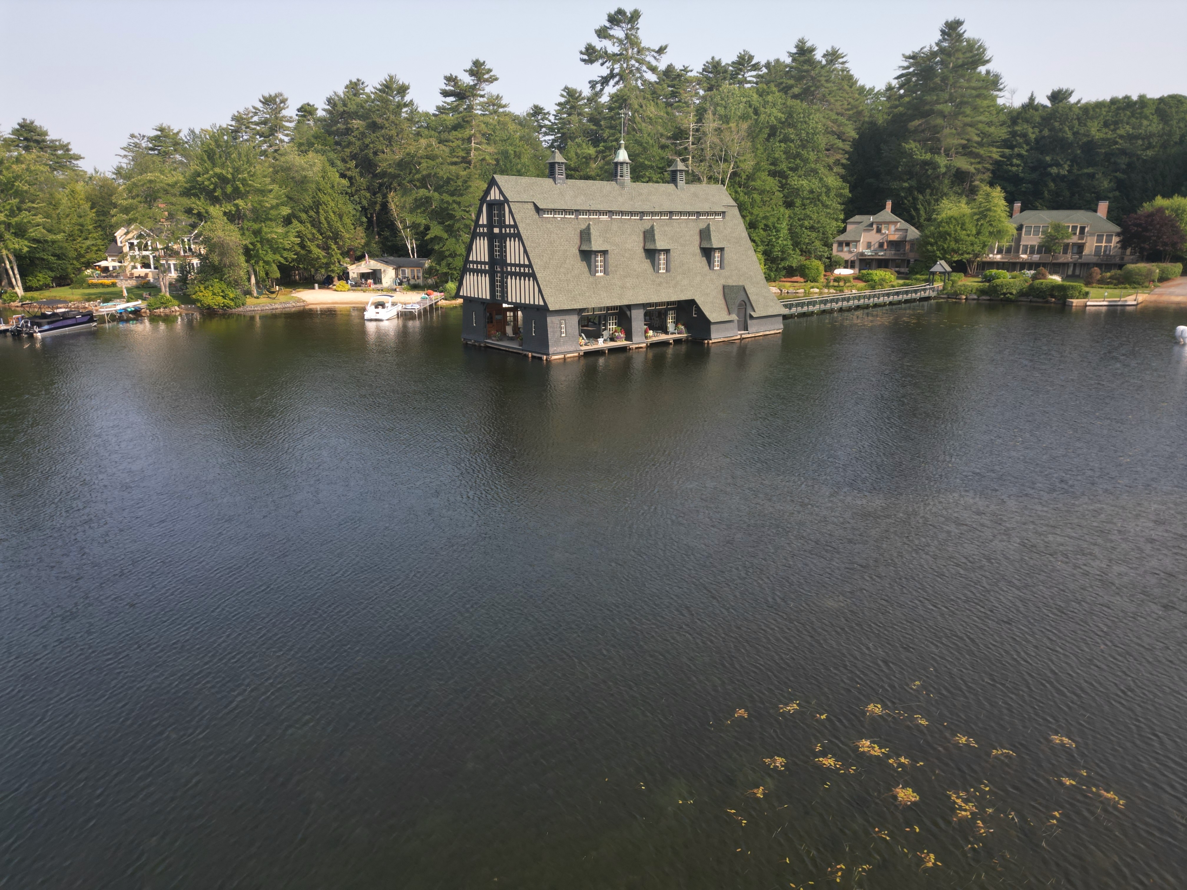 Aerial drone view of Tudor-style boathouse on a lake surrounded by trees and waterfront properties